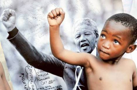 A young member of the Maitibolo Cultural Troupe, who came to dance for well-wishers in honour of Nelson Mandela, poses for well-wishers in front of a placard of Mandela, outside the entrance to the Mediclinic Heart Hospital where former South African President Nelson Mandela is being treated in Pretoria, South Africa Photo: AP